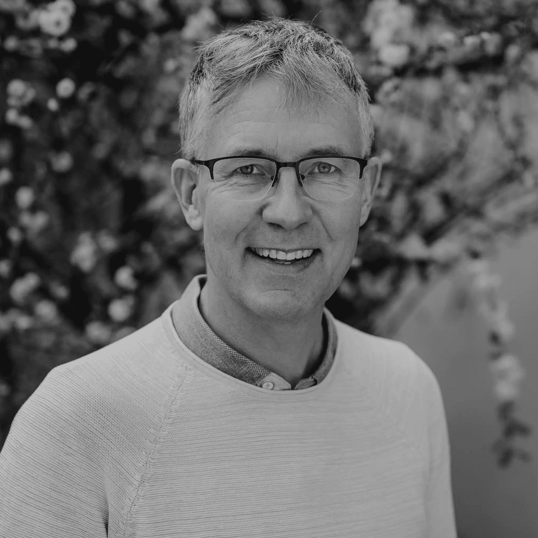 Gråhårig man med glasögon, skjorta och tröja tittar in i kameran och ler.