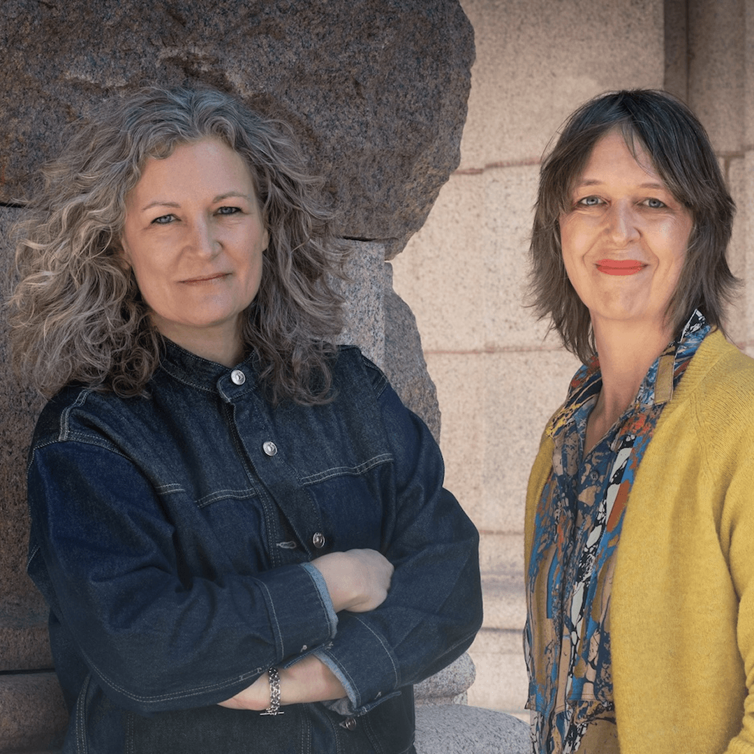 Två kvinnor. En med grått hår och en med brunt, poserar framför kameran.