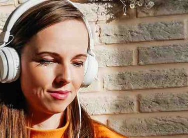 Bild på ung kvinna med hörlurar som lyssnar, mot en tegelvägg