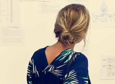 Vit kvinna med blont hår uppsatt i knut står med ryggen mot kameran och tittar på en vägg med texter. 