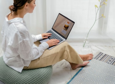 En vit kvinna sitter på en kudde på golvet med en dator i famnen. 